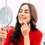Smiling brunette woman holding a mirror to view her Botox treatment.